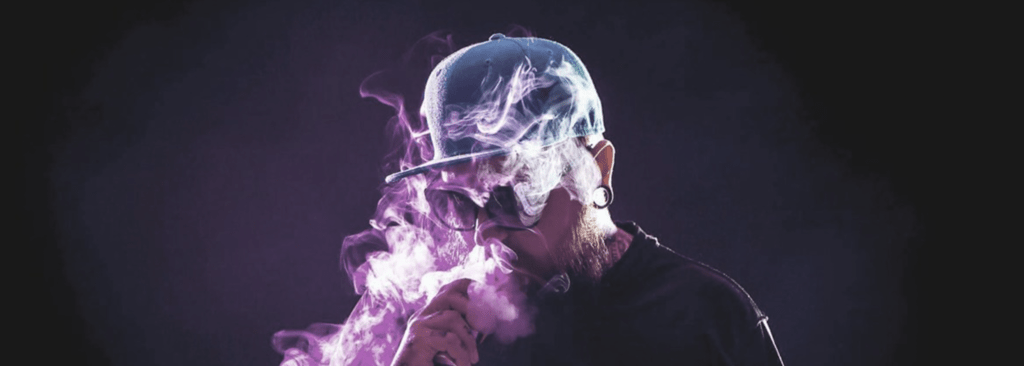 Un homme portant un chapeau soufflant de la fumée d'une cigarette électronique.