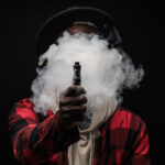 Une personne vêtue d'une chemise à carreaux et d'un chapeau exhalant un gros nuage de vapeur, masquant son visage et tenant une cigarette électronique dirigée vers la caméra.